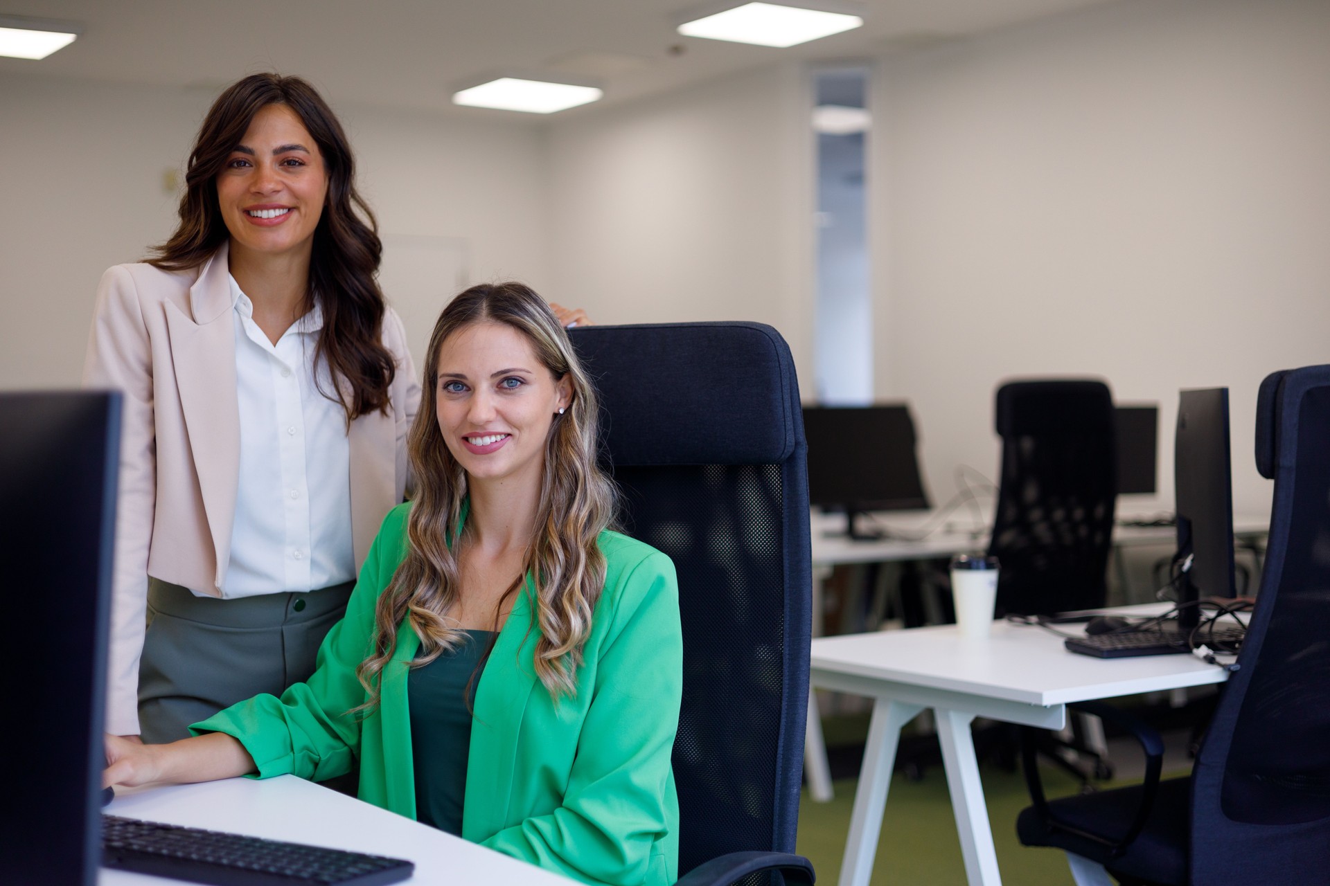 Two businesswomen smiling while collaborating at a computer in a modern office, showcasing teamwork and professionalism in their workspace