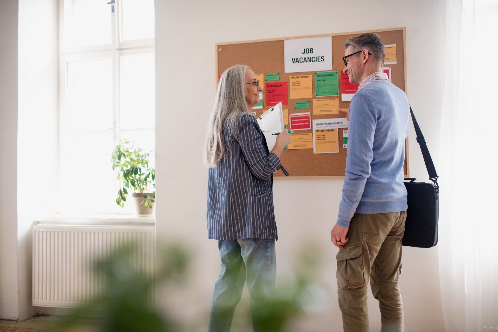 Recruitment agency employee standing in front of employment noticeboard and helping mature man search for a job.